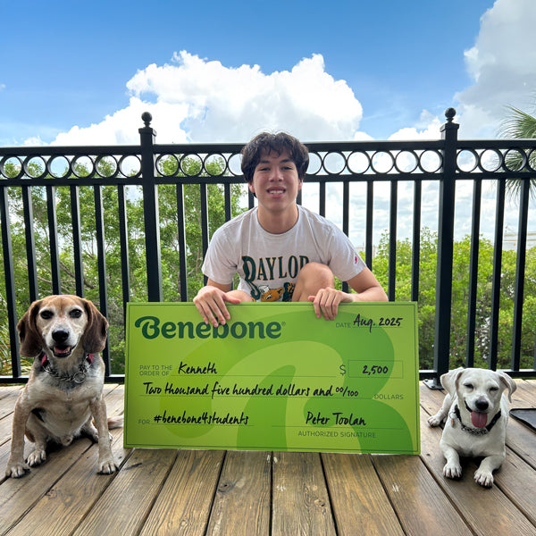 Benebone scholarship winner Kenneth holding a large green check, sitting outdoors with two dogs.