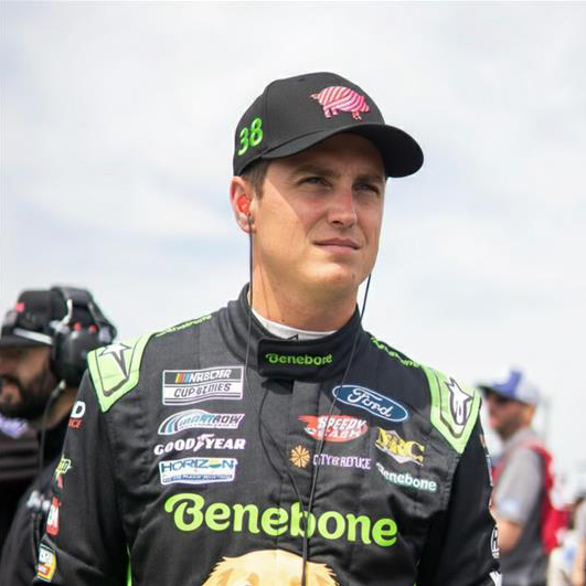 A racecar driver in a black racing suit with Benebone logos stands outdoors, wearing earphones and a Benebone Pig Hat. He looks ahead, showcasing his focus and pride in his sponsor and the unique hat.