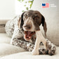 A brown and white spotted dog chews on a Benebone Green Wishbone while lying on a beige couch. A stylized American flag and “USA Made” appear in the top right corner, highlighting Benebone’s American-made Green Wishbone.
